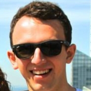 A smiling person wearing black sunglasses outdoors with a blue sky in the background.