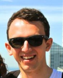 A smiling person wearing black sunglasses outdoors with a blue sky in the background.