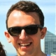 A smiling person wearing black sunglasses outdoors with a blue sky in the background.