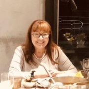 Smiling woman with glasses sits at a table with dishes, wearing a pearl necklace and light top.