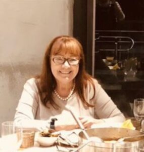 Smiling woman with glasses sits at a table with dishes, wearing a pearl necklace and light top.