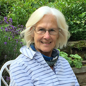 Smiling older woman with white hair, glasses, and striped shirt sitting outdoors among green plants.