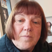 Older woman with short brown hair and bangs, wearing a dark jacket, looking directly at the camera.