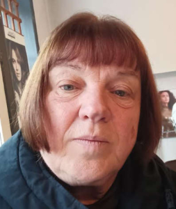 Older woman with short brown hair and bangs, wearing a dark jacket, looking directly at the camera.