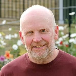 Bald, bearded man in a maroon shirt smiling outdoors with flowers and greenery in the background.