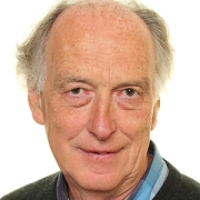 Older man with gray hair, wearing a collared shirt and sweater, smiling at the camera against a white background.