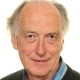 Older man with gray hair, wearing a collared shirt and sweater, smiling at the camera against a white background.