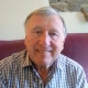 An older man with gray hair smiles while sitting on a maroon couch, wearing a plaid shirt.