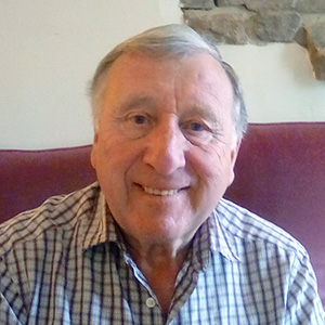 An older man with gray hair smiles while sitting on a maroon couch, wearing a plaid shirt.
