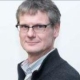 A man with short gray hair and glasses, wearing a dark sweater over a collared shirt, looks at the camera.