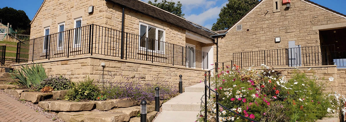 A stone house with a sloped roof, garden, and pathway under a blue sky with scattered clouds.