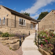 A stone house with a sloped roof, garden, and pathway under a blue sky with scattered clouds.