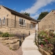 A stone house with a sloped roof, garden, and pathway under a blue sky with scattered clouds.